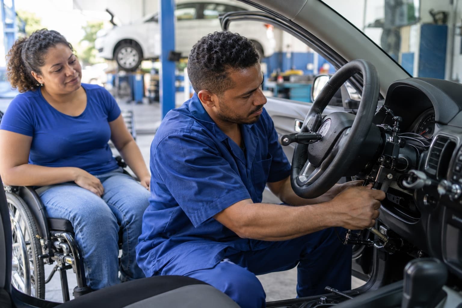 Técnico de uniforme azul instalando comandos manuais de acelerador e freio na coluna de direção de um carro, com um cadeirante acompanhando ao lado da porta aberta