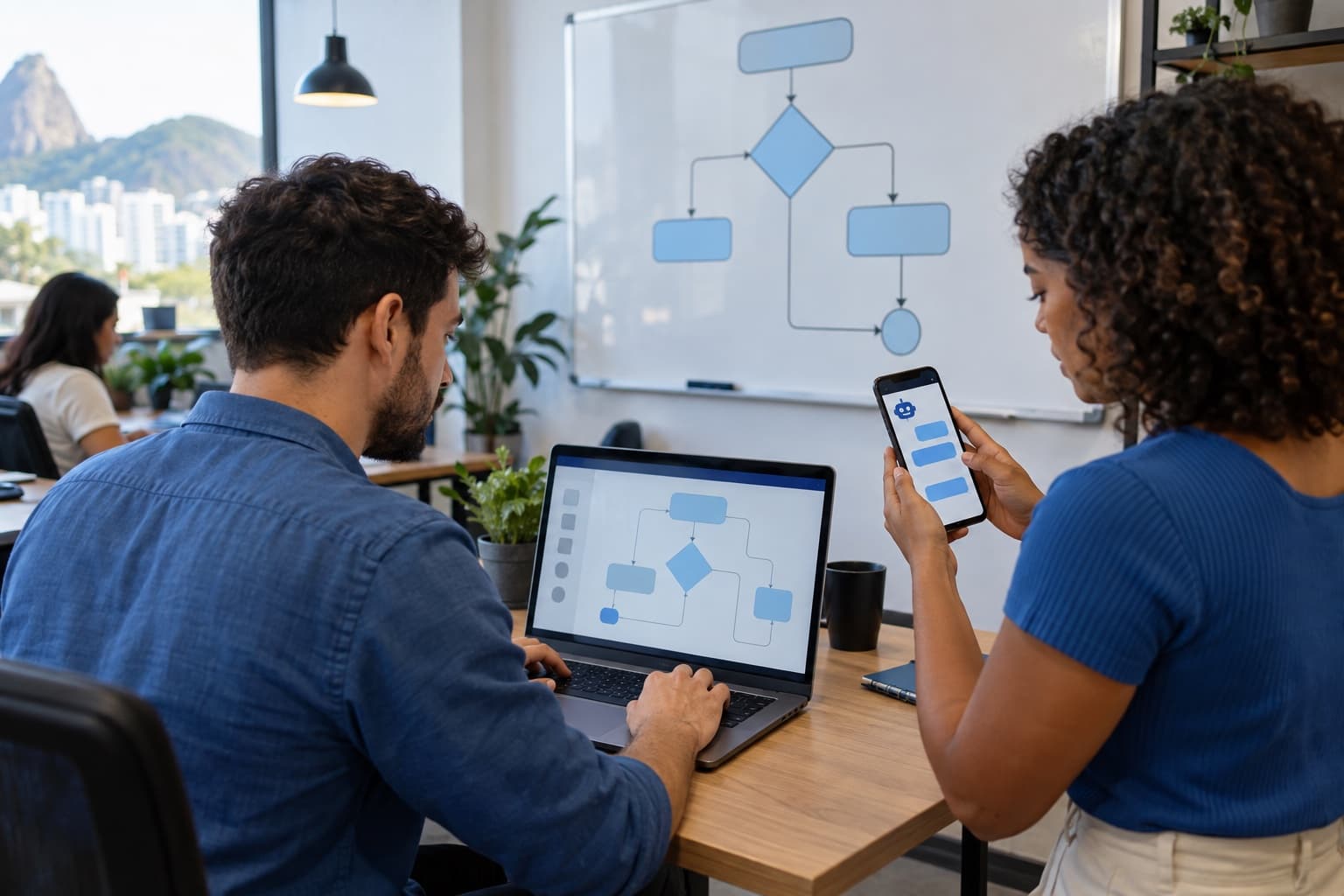 Desenvolvedor de camisa azul montando um fluxo de atendimento num notebook enquanto o colega testa no celular, com um quadro branco de fluxograma ao fundo