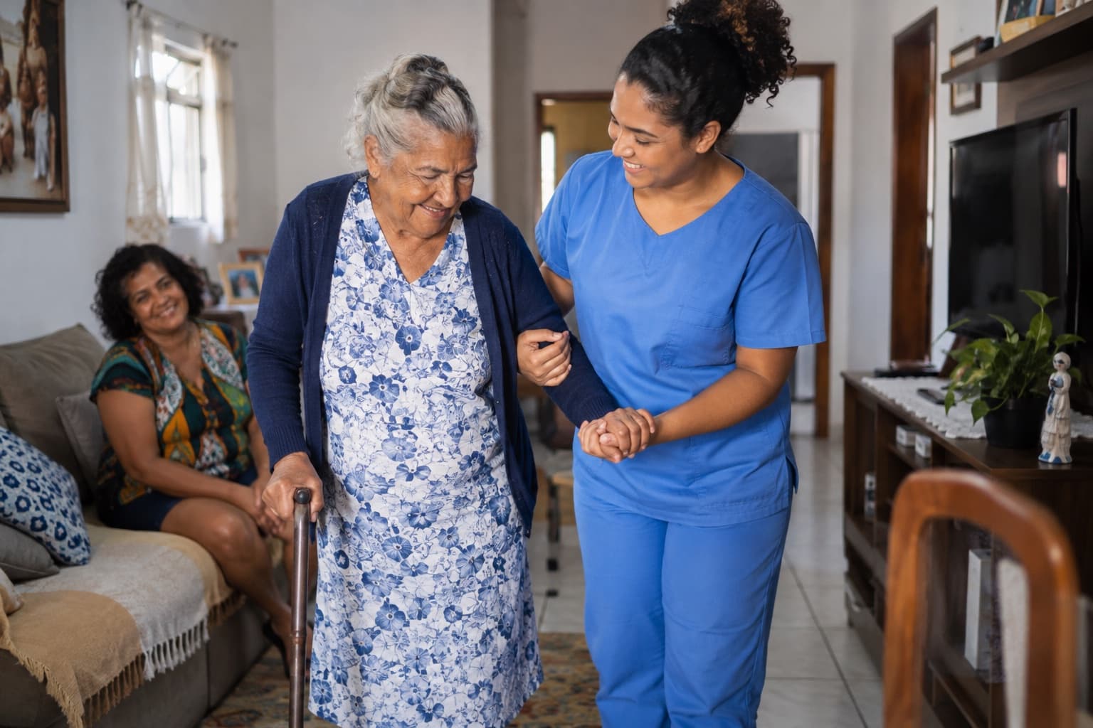 Cuidadora de uniforme azul ajudando uma senhora idosa a caminhar na sala de casa enquanto uma familiar observa sorrindo