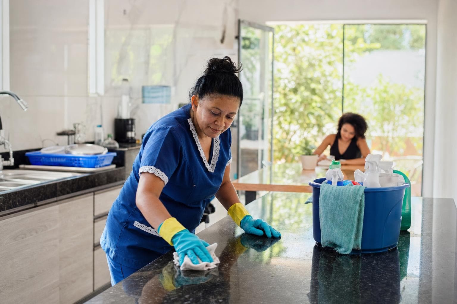 Diarista de uniforme azul limpando a bancada da cozinha de uma casa iluminada