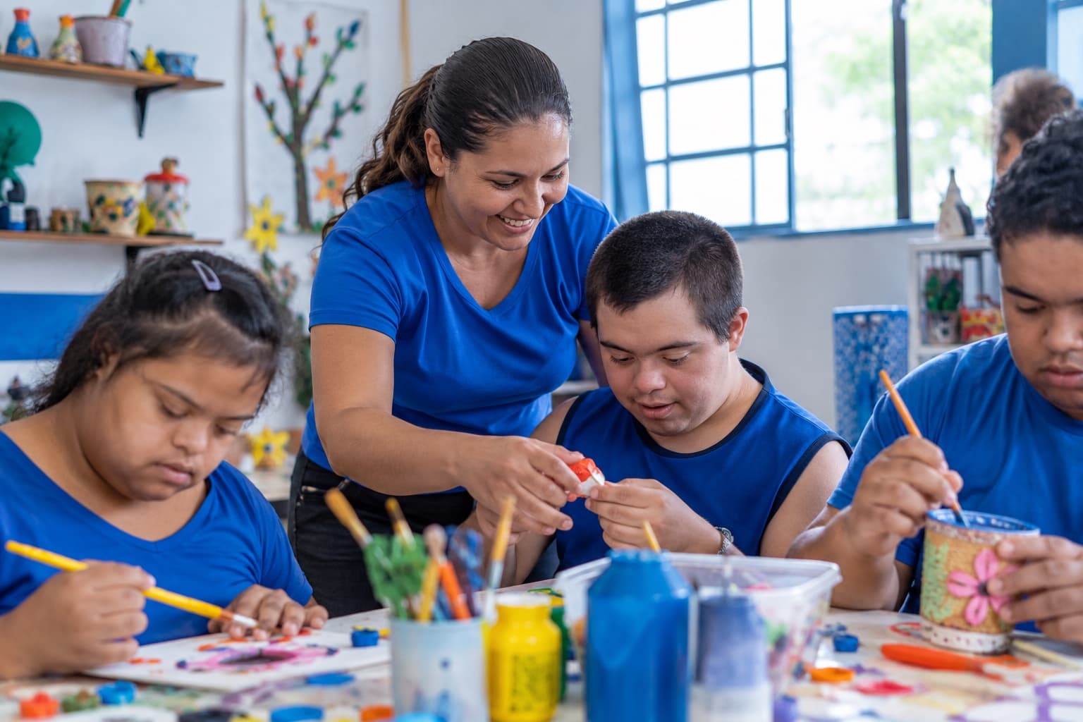 Terapeuta de camisa azul ajudando jovens com deficiência intelectual numa oficina de artesanato, com materiais coloridos sobre as mesas e luz natural
