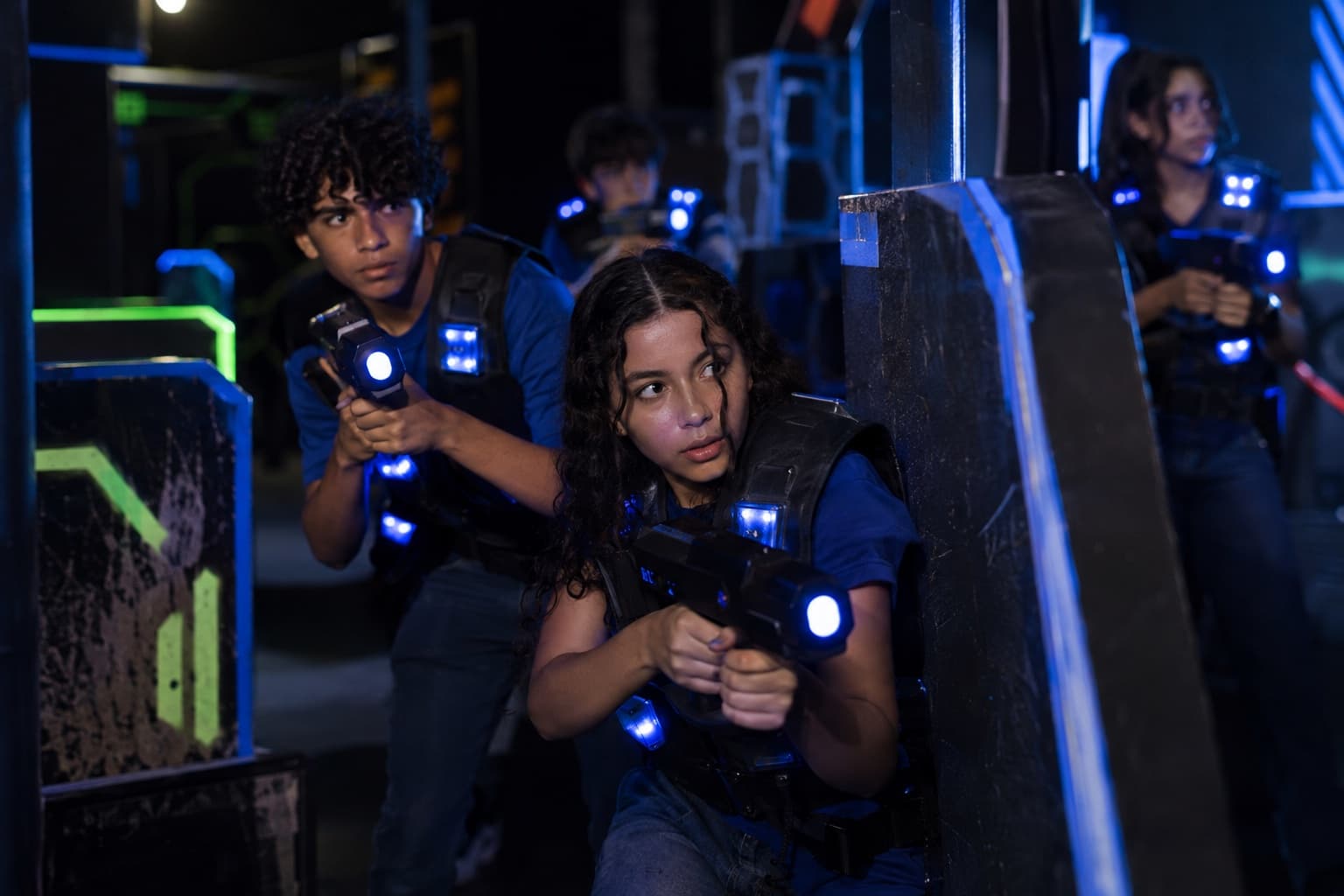 Adolescentes com coletes de laser tag e pistolas de laser em uma arena escura com obstáculos iluminados em neon, no meio de uma partida