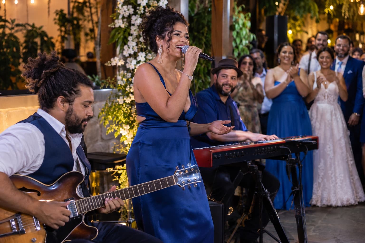 Banda ao vivo com vocalista, guitarrista e tecladista tocando em uma recepção de casamento, com convidados batendo palmas e luz quente de palco