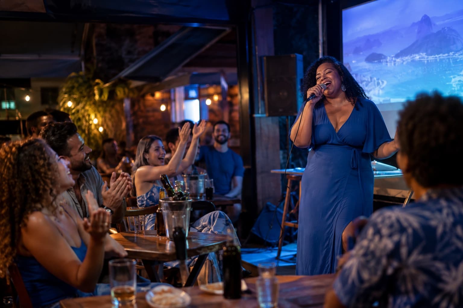 Bar de karaokê à noite, uma mulher cantando ao microfone em um pequeno palco enquanto amigos nas mesas comemoram, com o brilho de uma tela ao fundo