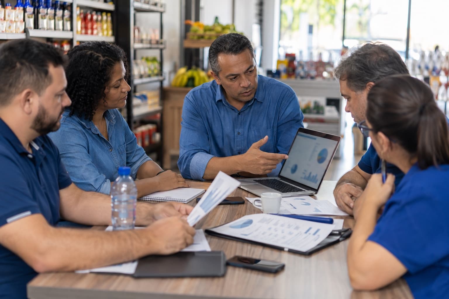 Donos de pequenos supermercados reunidos numa mesa comparando cotações de fornecedores no notebook e em folhas impressas, com um coordenador de camisa azul apresentando
