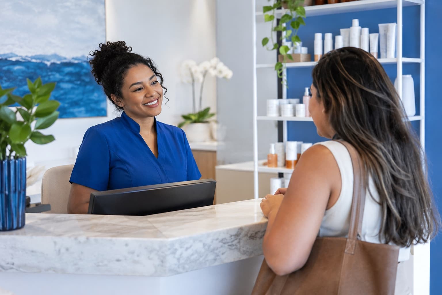 Recepcionista de uniforme azul atendendo uma cliente no balcão de uma clínica de estética clara com prateleiras de dermocosméticos