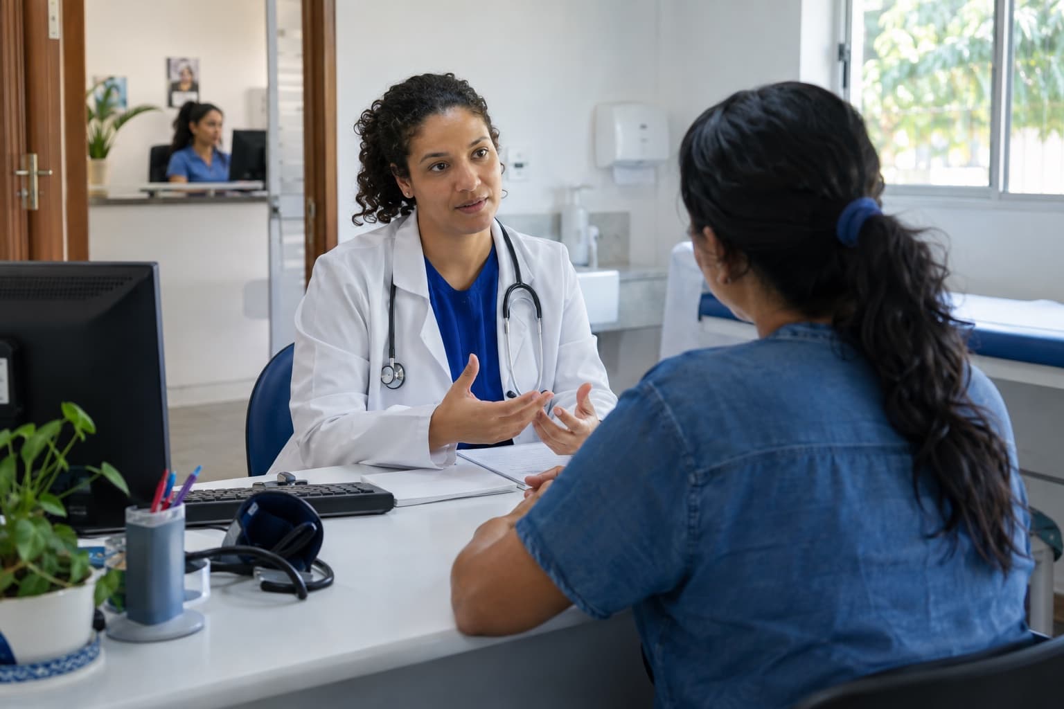 Médico de jaleco branco conversando com um paciente na mesa do consultório, com a recepcionista ao computador vista pela porta aberta