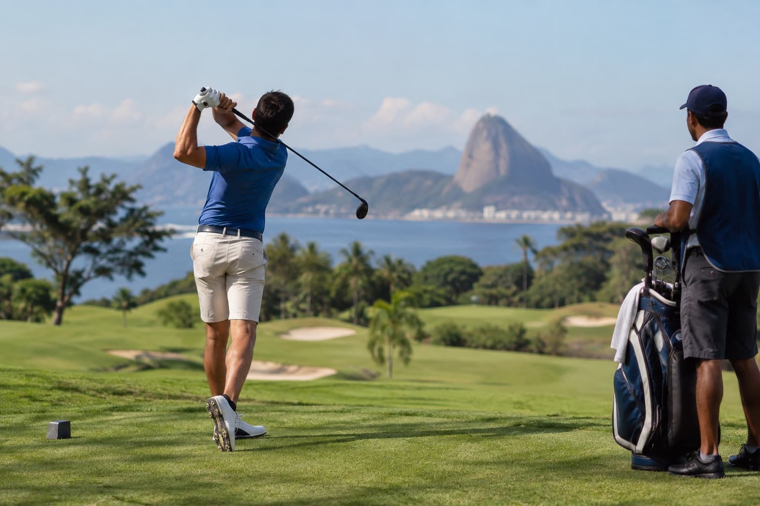 Golfista de camisa polo azul dando a tacada de saída em um fairway verde de um clube de golfe, com o caddie ao lado segurando a bolsa de tacos