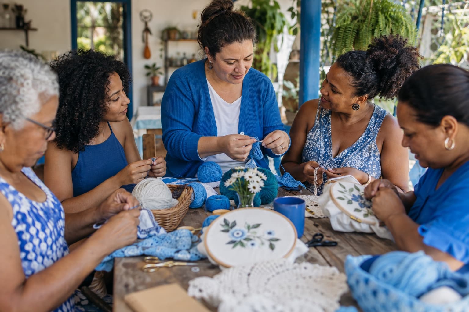 Grupo de mulheres de várias idades ao redor de uma mesa de madeira aprendendo crochê e bordado com uma instrutora de cardigã azul