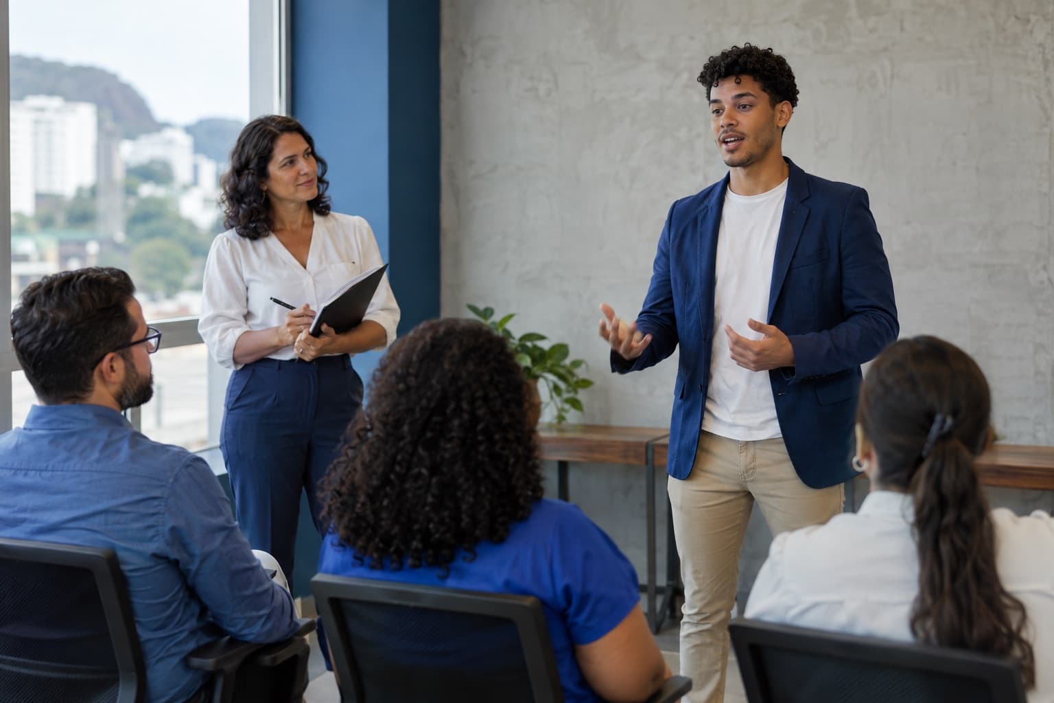 Jovem de blazer azul praticando apresentação em pé diante de um pequeno grupo sentado numa sala de treinamento, com a instrutora observando