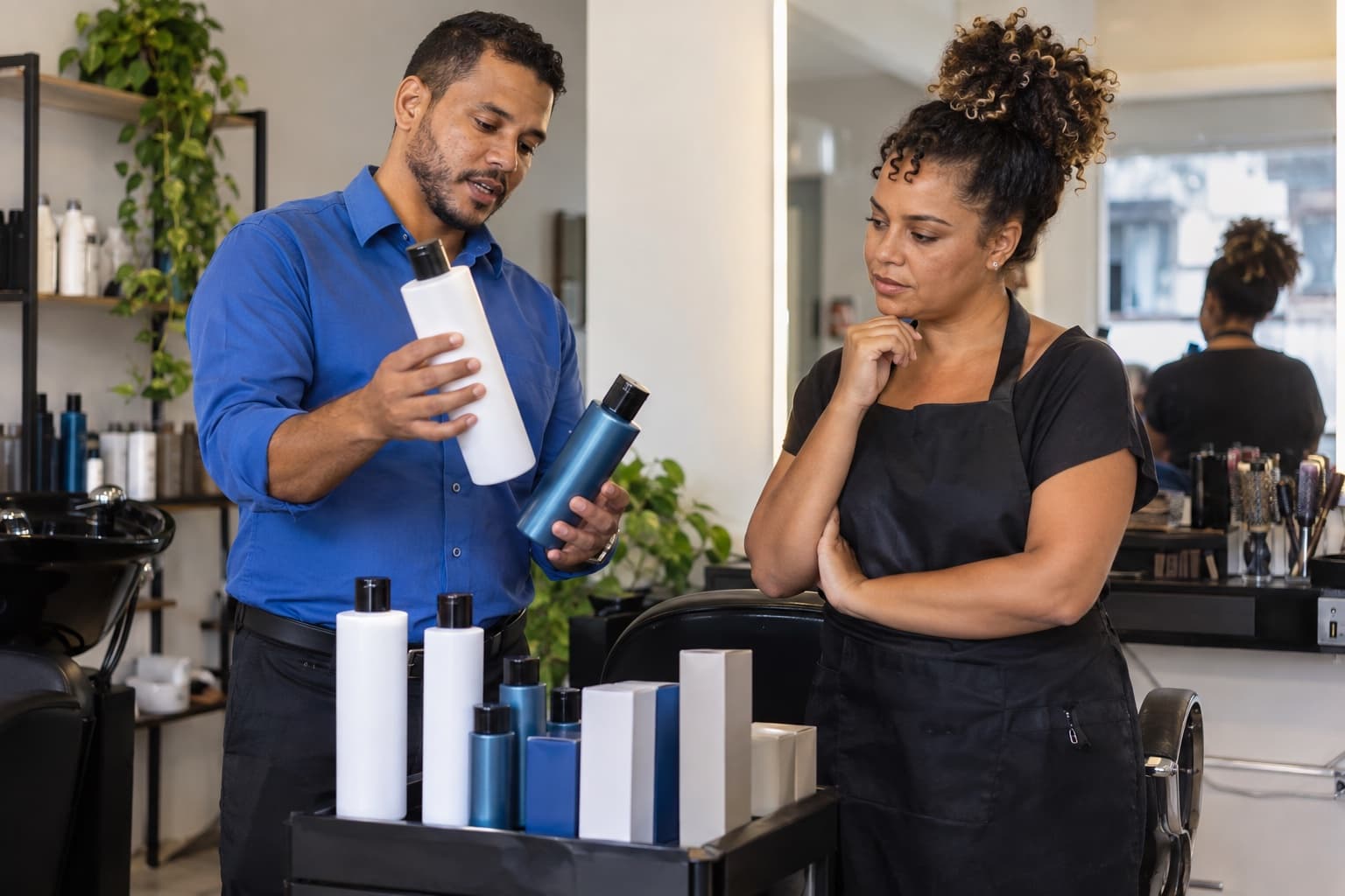 Vendedor de camisa azul mostrando frascos de tratamento capilar e caixas de coloração a uma cabeleireira dentro de um salão de beleza