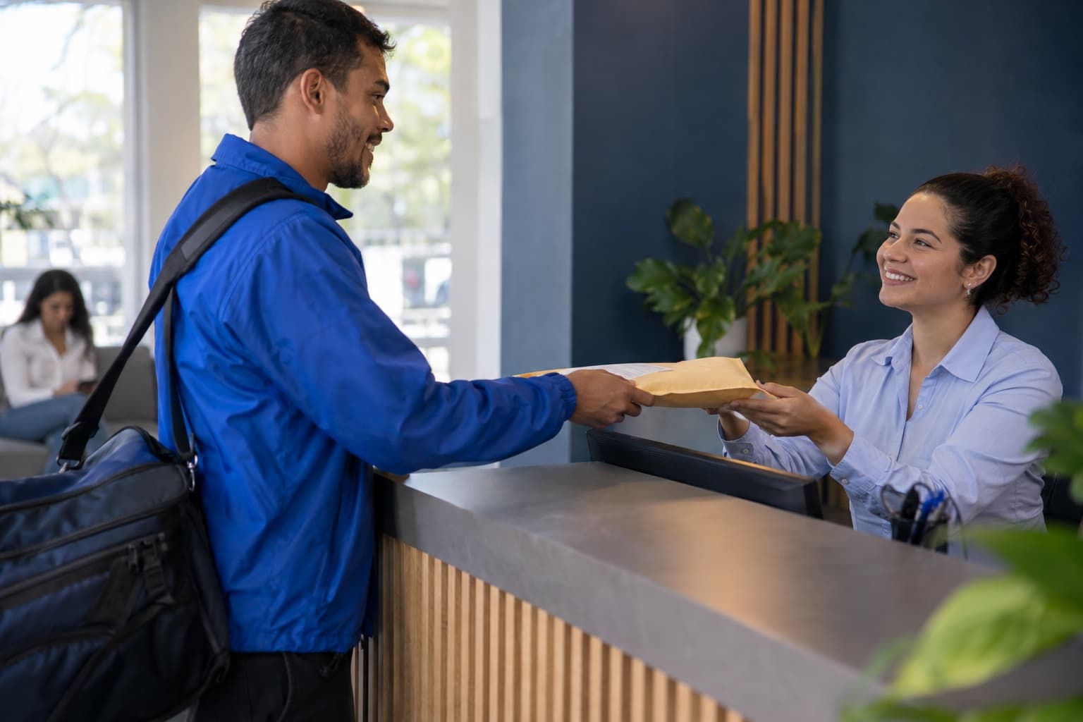 Entregador de jaqueta azul entregando envelope lacrado à recepcionista no balcão de um escritório, com bolsa de entregas no ombro