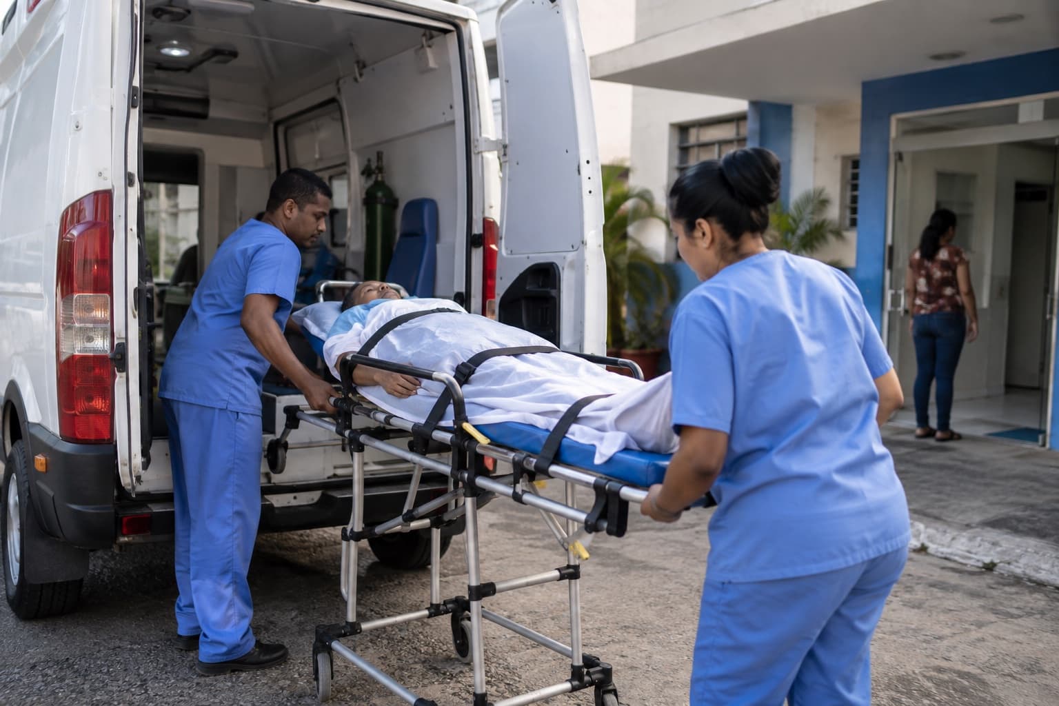 Dois técnicos de enfermagem de uniforme azul colocando paciente em maca na traseira de uma ambulância branca estacionada em frente a um hospital