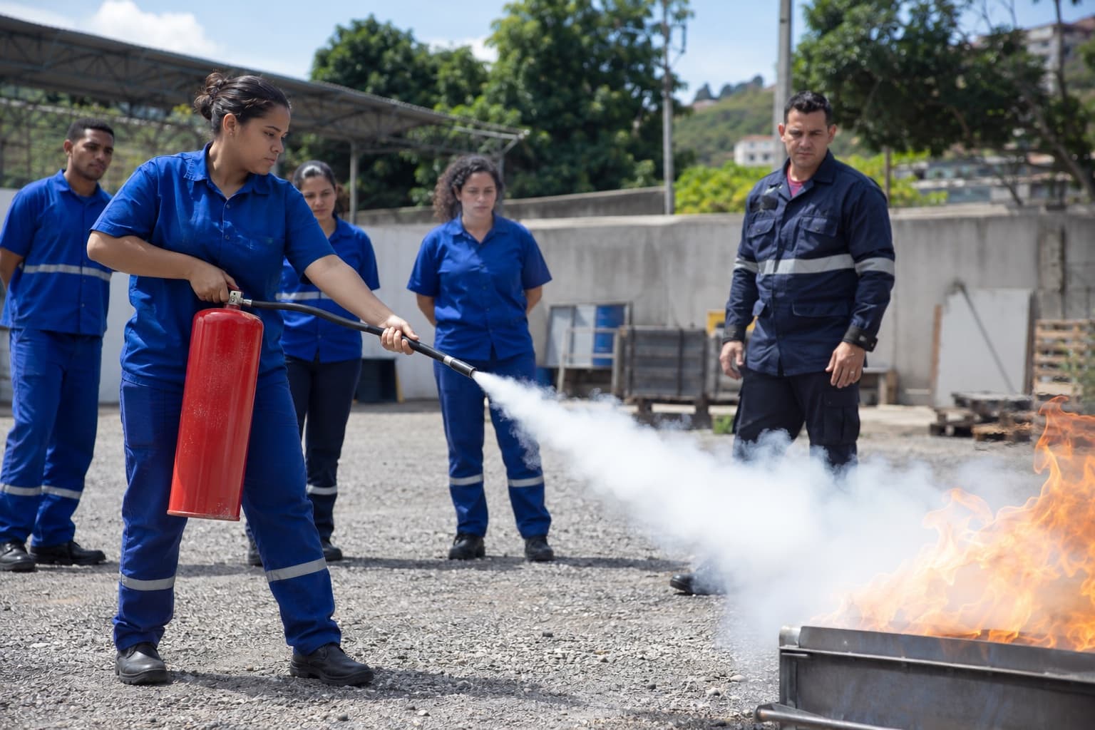 Alunos de curso de bombeiro civil de uniforme azul apagando um fogo controlado com extintor em um pátio de treinamento enquanto o instrutor observa