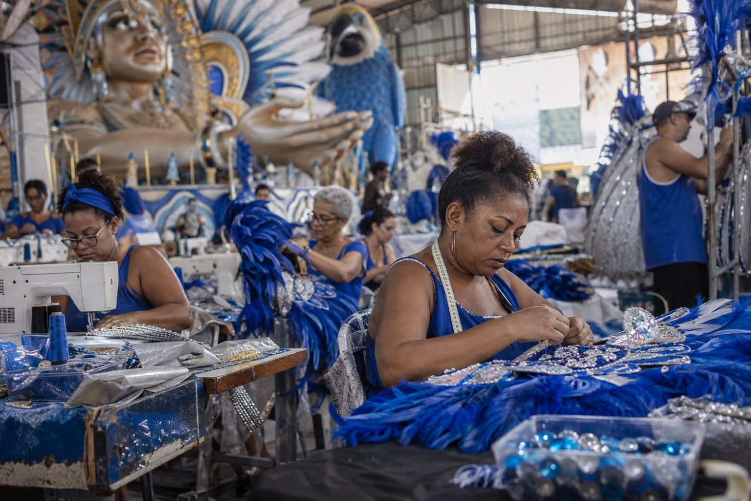 Costureiras de azul bordando fantasias com plumas e pedrarias num barracão de escola de samba, com parte de um carro alegórico ao fundo