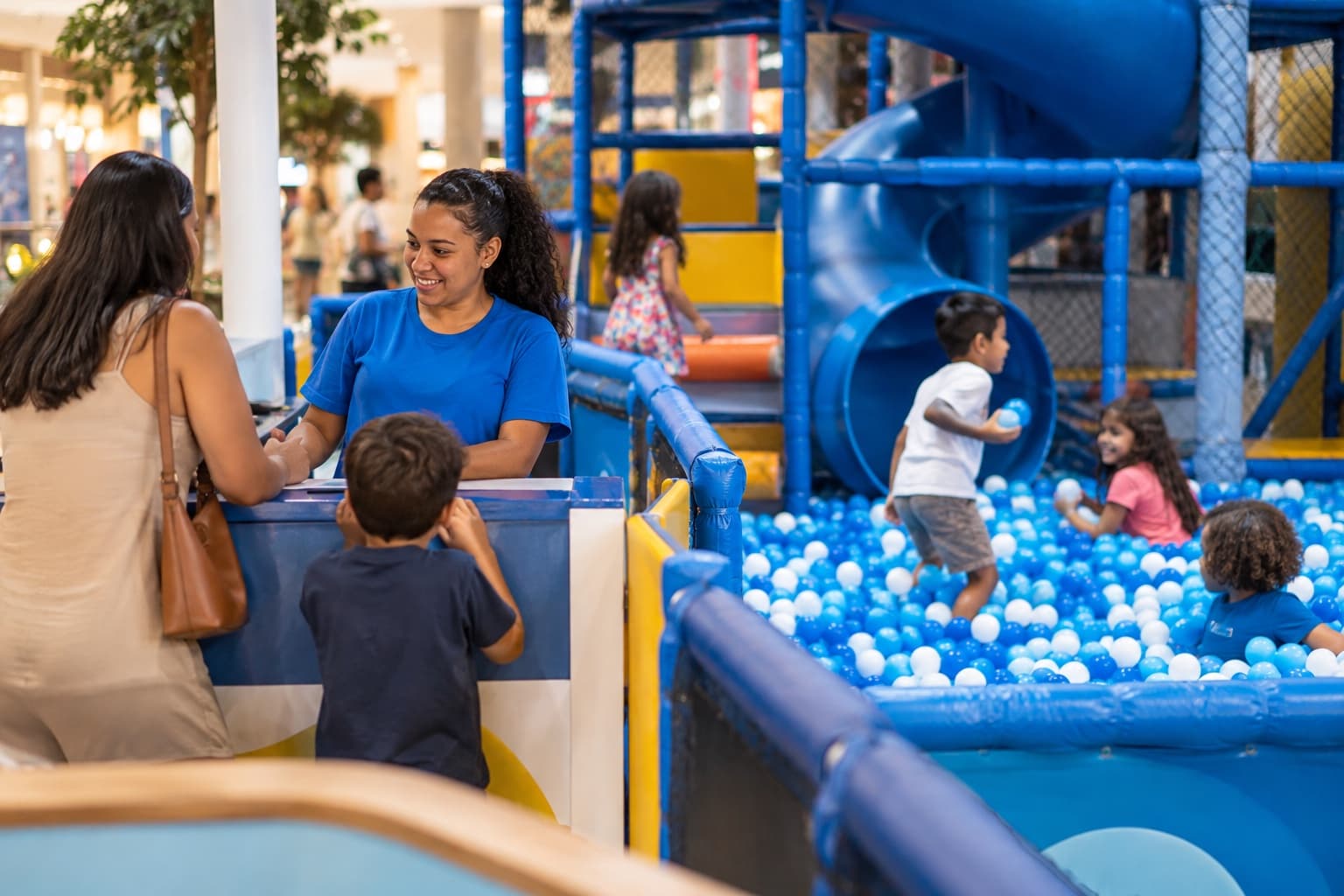 Área de brincar infantil em shopping com piscina de bolinhas e playground macio, crianças brincando enquanto uma monitora de camiseta azul acompanha e um pai faz o check-in no balcão