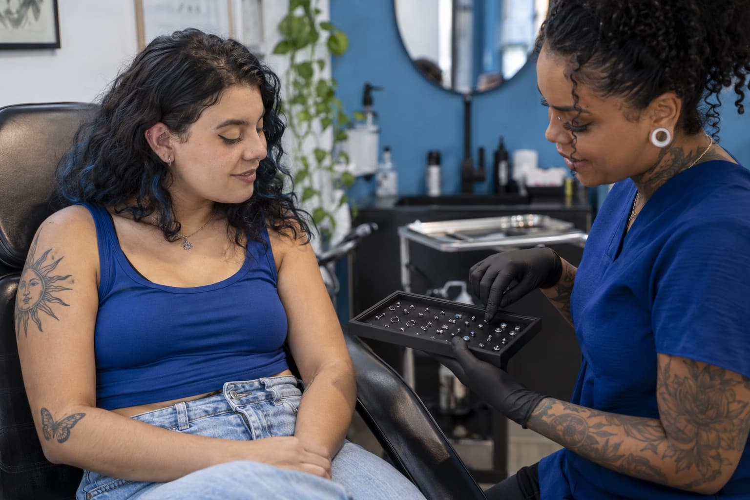 Body piercer de luvas pretas mostrando uma bandeja de joias de titânio a uma cliente sentada na cadeira do estúdio