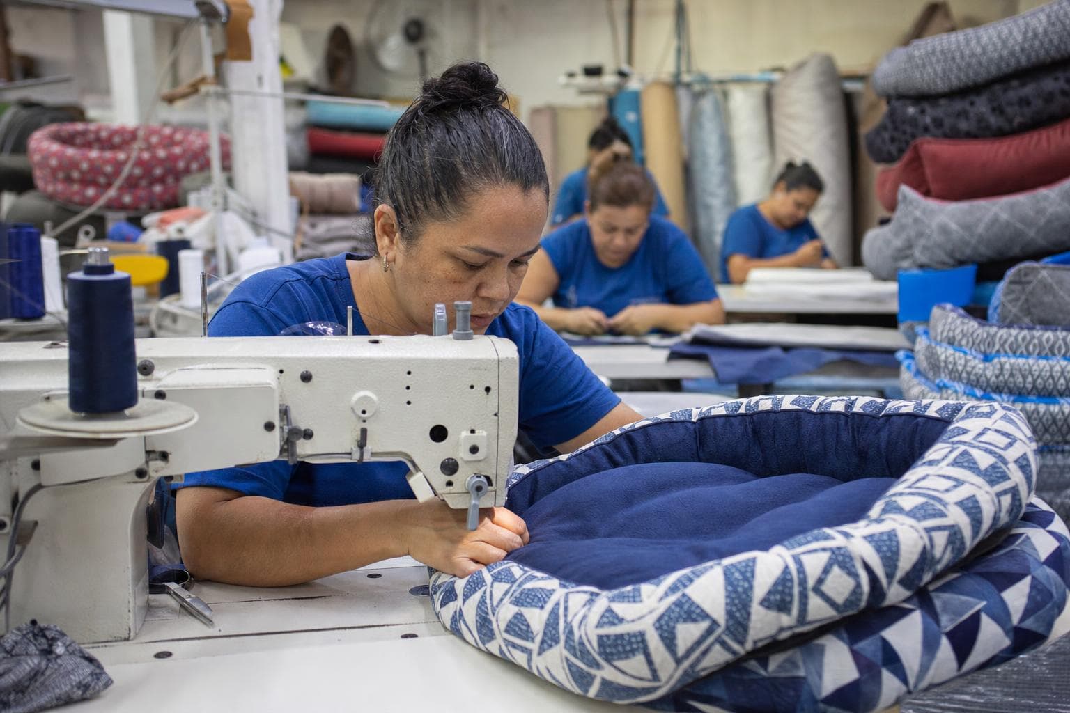 Costureira fazendo uma caminha acolchoada para cães em máquina industrial, com caminhas empilhadas ao fundo