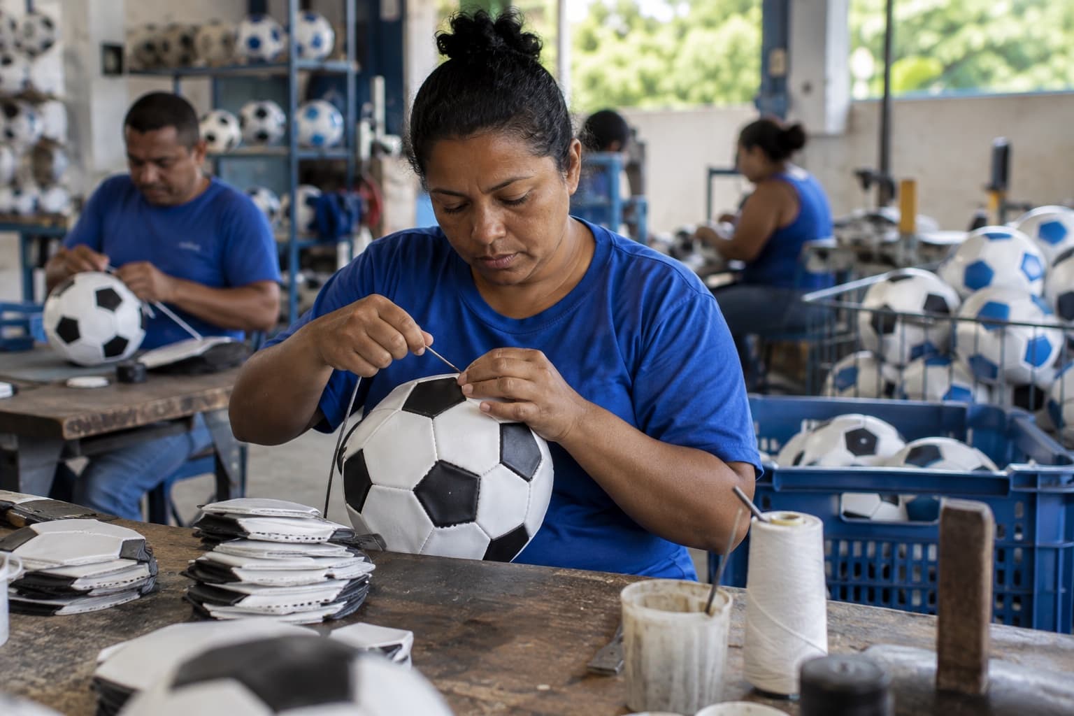Trabalhador de camiseta azul costurando à mão os gomos de uma bola de futebol em uma bancada, com bolas prontas em cestos na fábrica