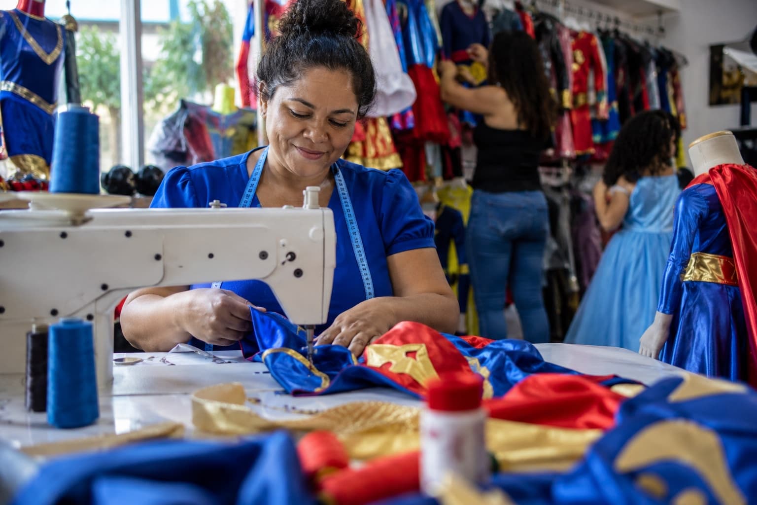 Costureira de blusa azul costurando uma fantasia infantil de super-herói colorida numa oficina de fantasias, com araras cheias de fantasias ao fundo