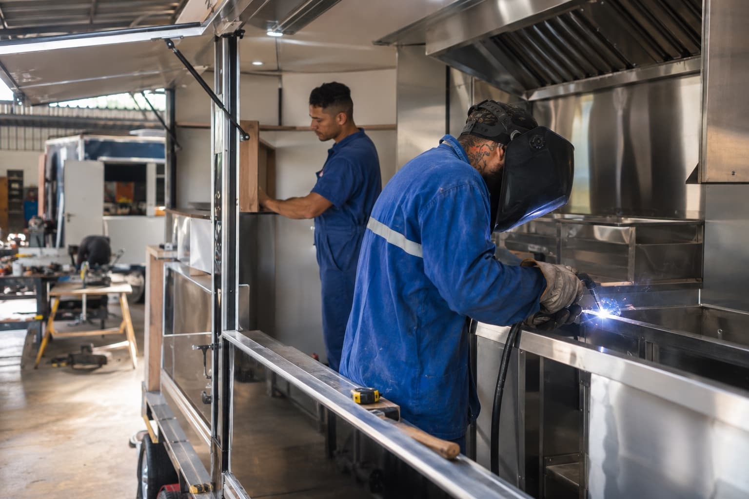 Soldador e marceneiro de macacão azul montando o interior de um trailer de food truck na oficina, instalando bancadas de inox