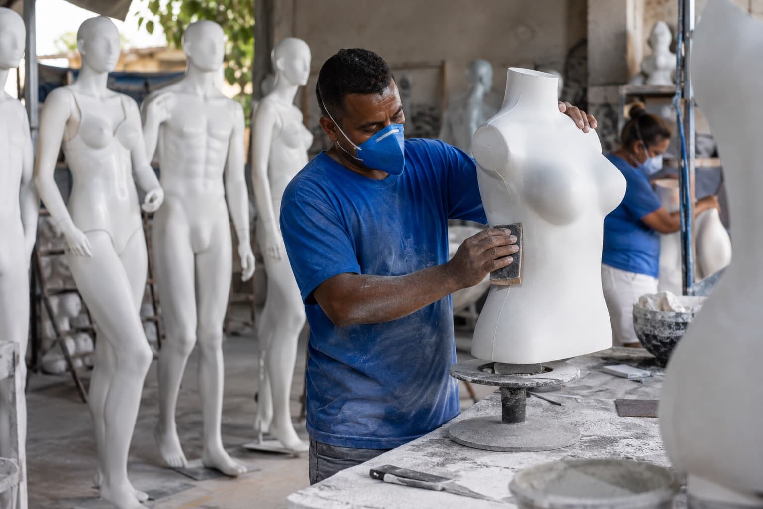 Funcionário de camisa azul e máscara lixando um torso de manequim de fibra branco numa pequena fábrica, com manequins prontos enfileirados ao fundo