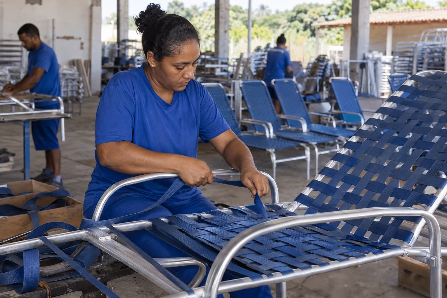 Funcionária de uniforme azul trançando fita sintética numa espreguiçadeira de alumínio numa pequena fábrica, com cadeiras de piscina prontas ao fundo