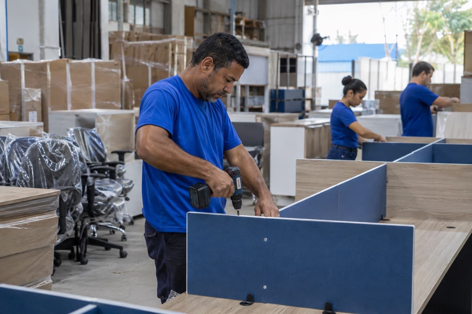 Montador de camisa azul fixando divisórias em estações de trabalho de escritório dentro de uma fábrica de móveis, com mesas e cadeiras embaladas ao lado