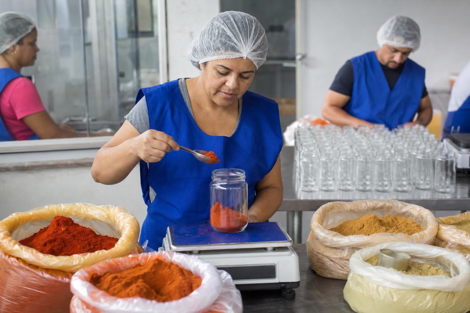 Trabalhador pesando páprica em balança digital ao lado de sacos de especiarias em uma fábrica de temperos