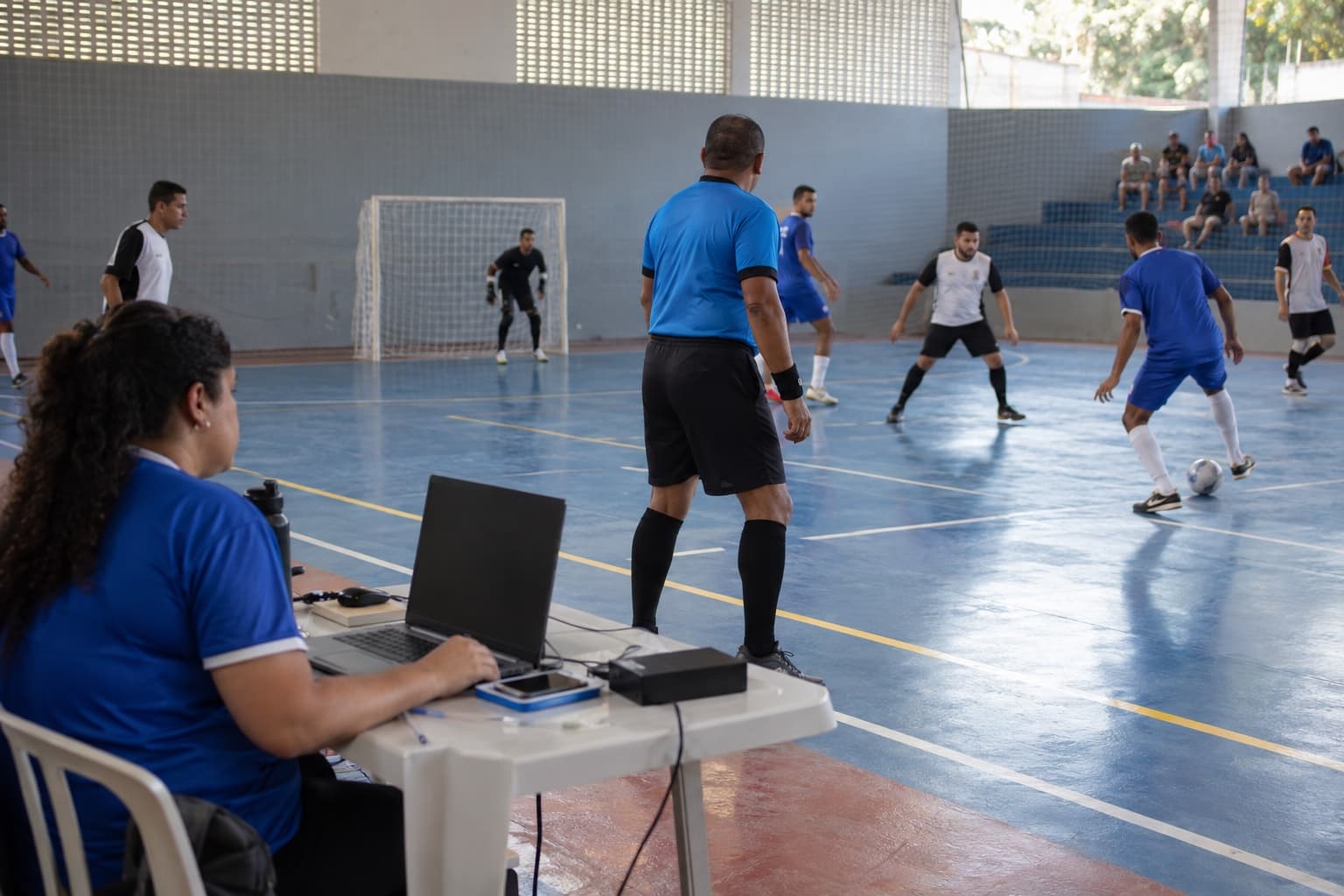 Partida de liga amadora de futsal em ginásio, árbitro de camisa azul e organizador na mesa com notebook