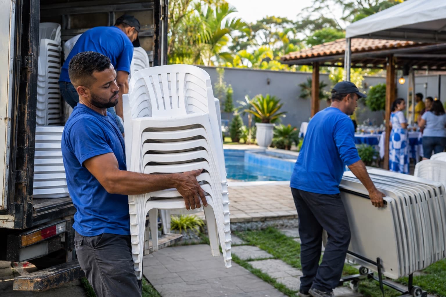 Funcionários descarregando cadeiras plásticas brancas empilhadas e mesas dobráveis de um caminhão num quintal de festa