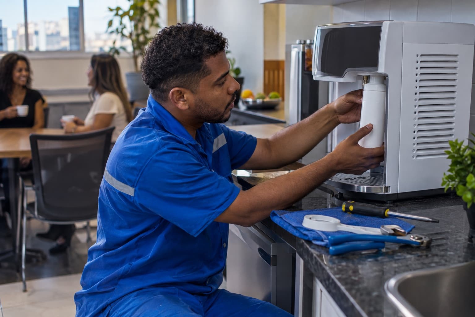 Técnico de uniforme azul instalando purificador de água na copa de um escritório, trocando o refil com ferramentas sobre a bancada
