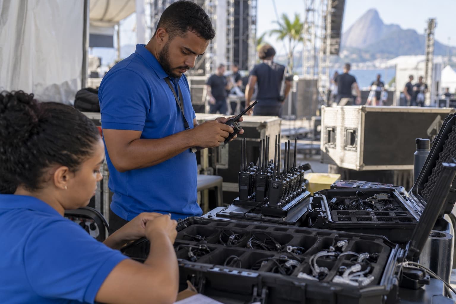 Funcionário de camisa azul testando rádios comunicadores num carregador múltiplo, com maletas de rádios e fones sobre a mesa nos bastidores de um evento