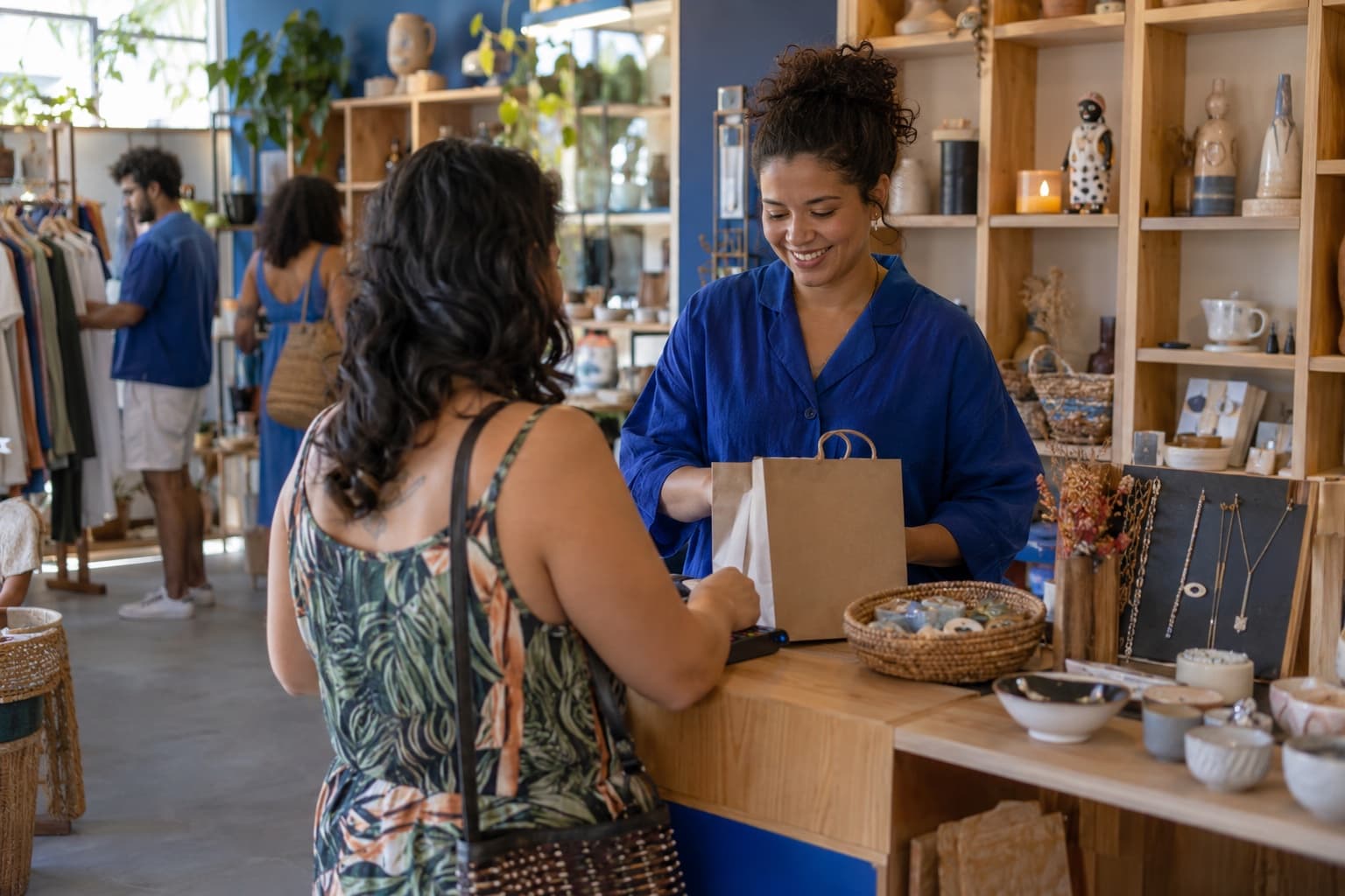 Atendente de blusa azul no caixa de uma loja colaborativa, com prateleiras e nichos de várias marcas locais exibindo cerâmica, velas, bijuterias e roupas