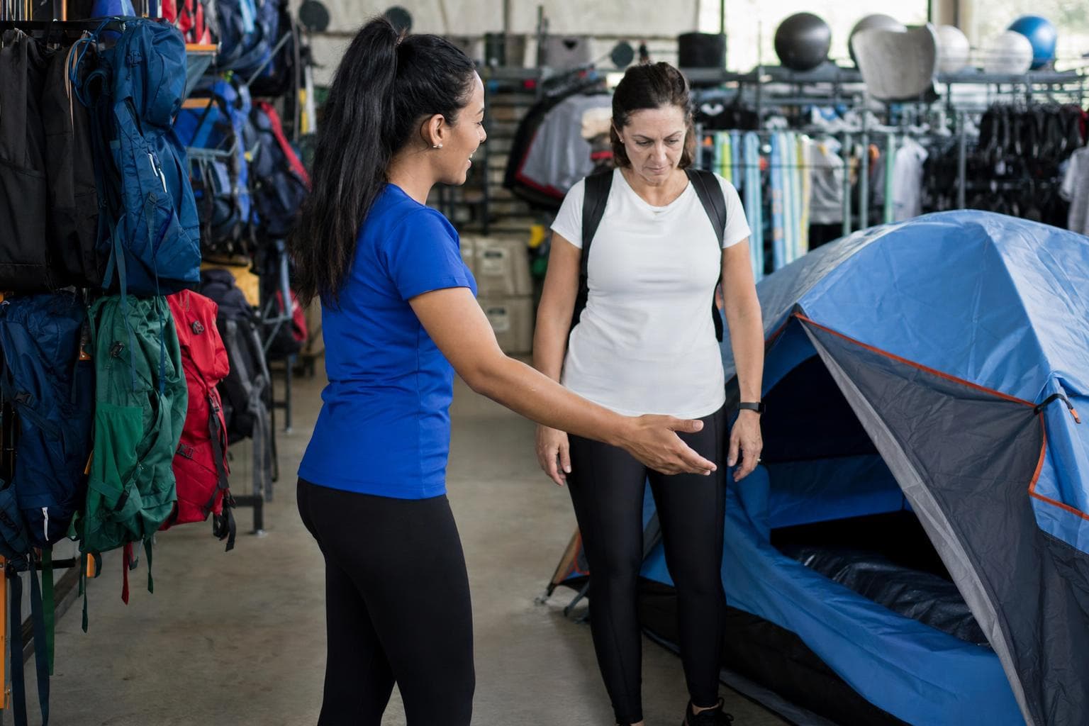 Vendedora mostrando uma barraca montada e mochilas a um cliente em uma loja de camping