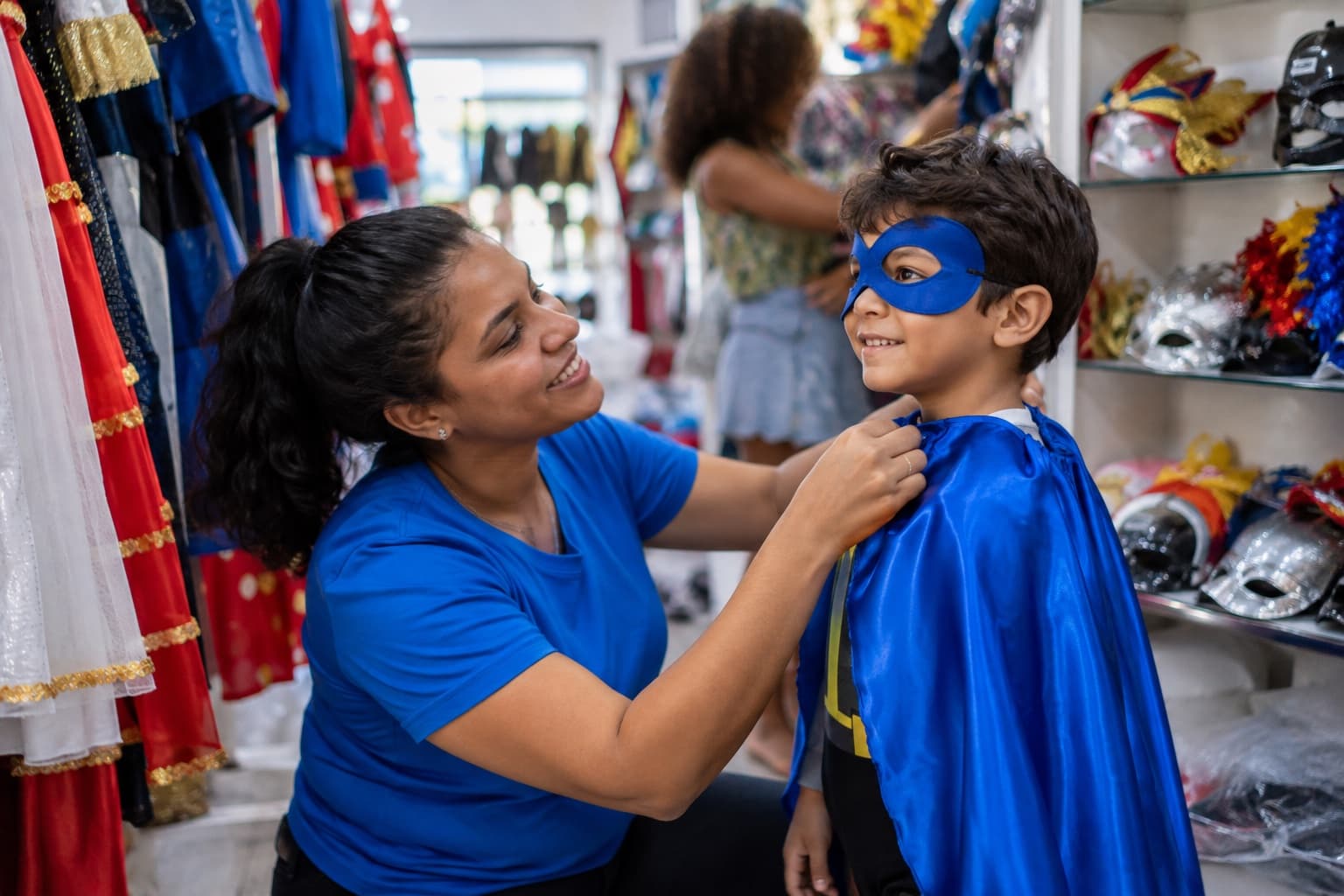 Loja com araras de fantasias coloridas de carnaval e festa, atendente de camisa azul ajudando uma criança a experimentar uma capa de super-herói, com máscaras nas prateleiras