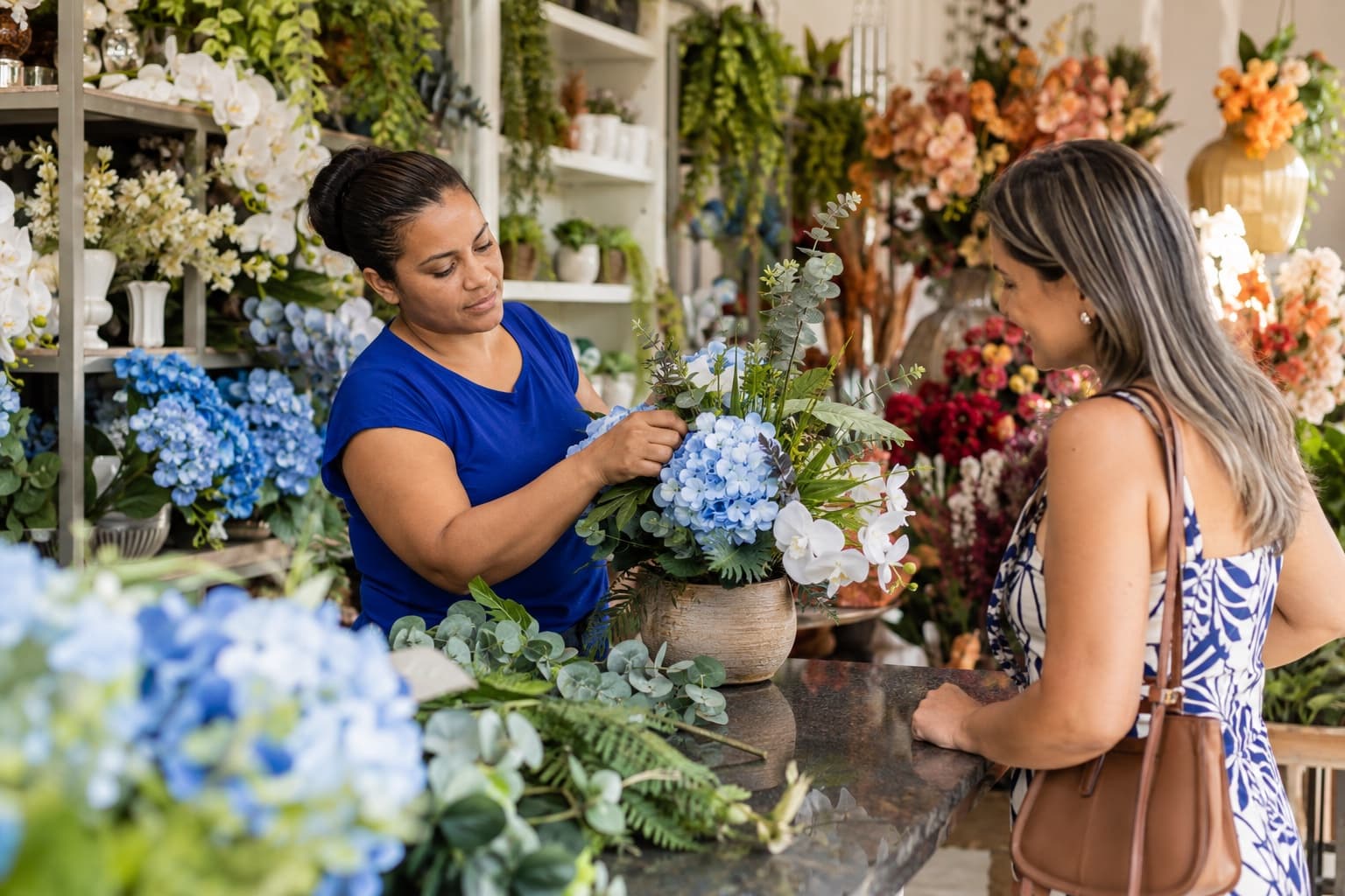 Vendedora de blusa azul montando um arranjo decorativo para uma cliente numa loja com paredes cheias de flores de seda, folhagens e plantas artificiais