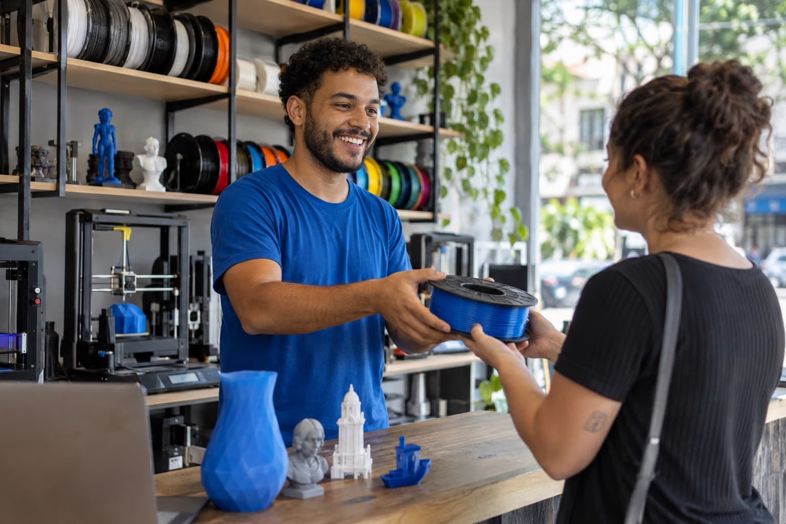 Atendente de camiseta azul entregando um carretel de filamento a uma cliente numa loja com prateleiras de filamentos coloridos e impressoras 3D em exposição