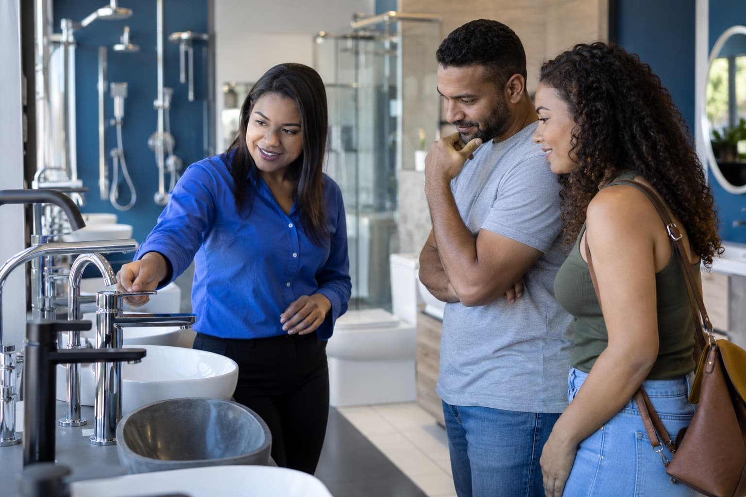 Vendedora de blusa azul mostrando um misturador monocomando a um casal num showroom de louças e metais com cubas, torneiras e chuveiros expostos