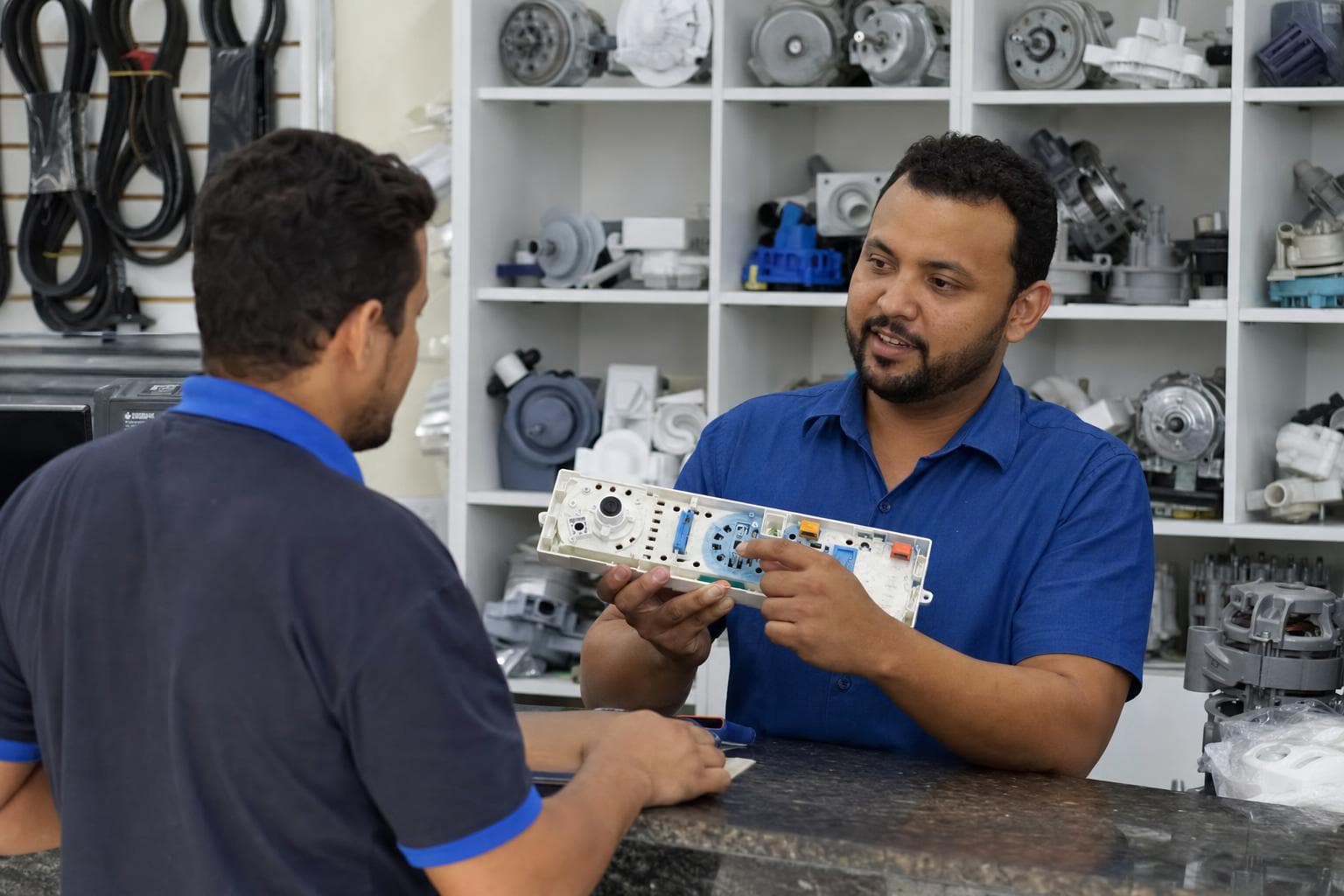 Balconista mostrando uma placa eletrônica de máquina de lavar a um técnico no balcão de uma loja de peças
