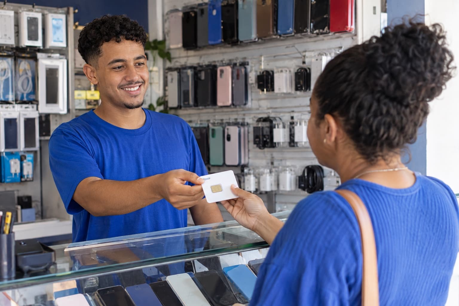 Atendente jovem de camiseta azul entregando um chip de celular a uma cliente no balcão de uma loja de acessórios para celular com capinhas e carregadores na parede