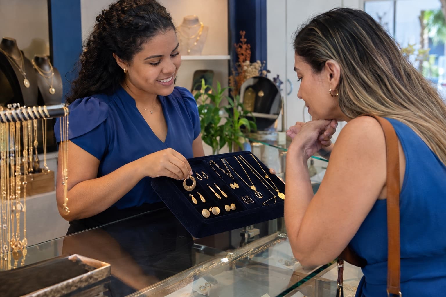 Vendedora de blusa azul mostrando brincos e colares folheados a ouro em uma bandeja de veludo para uma cliente no balcão de vidro de uma loja de semijoias