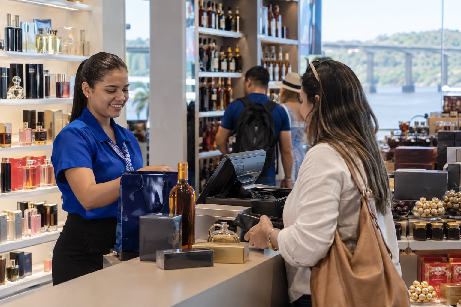 Vendedora de uniforme azul atendendo um viajante no caixa de um free shop de fronteira, com prateleiras de perfumes, bebidas e chocolates ao fundo