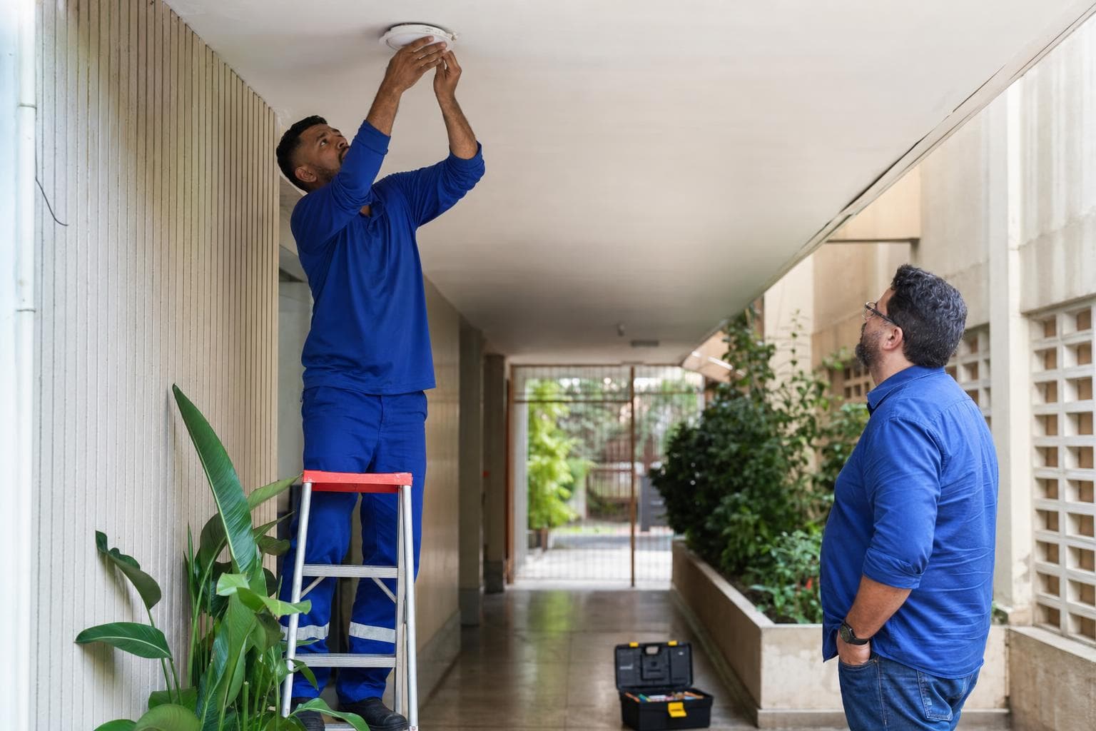 Técnico de manutenção em uniforme azul trocando uma luminária no corredor de um condomínio