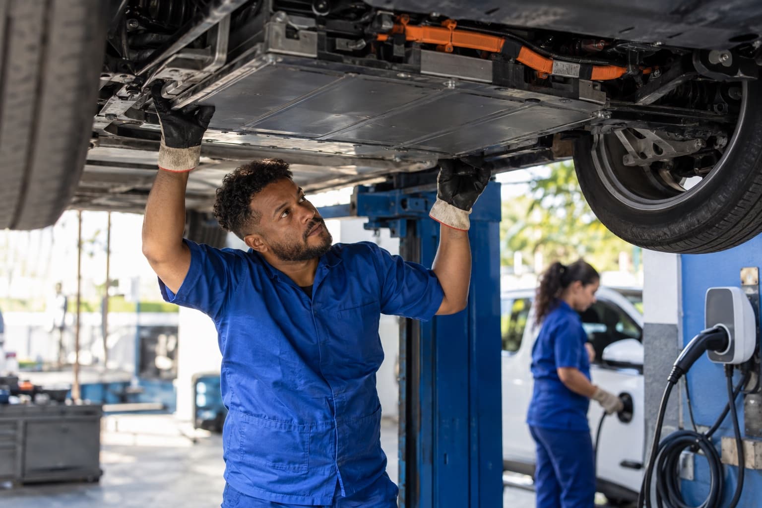 Mecânico de uniforme azul com luvas isolantes inspecionando a bateria sob um carro elétrico erguido no elevador, com cabo de recarga ao lado