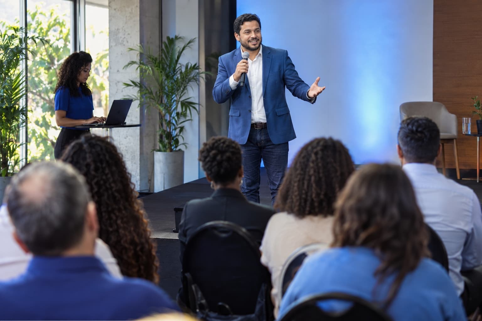 Palestrante de blazer azul falando em palco de evento corporativo, plateia de costas e assistente com notebook ao lado