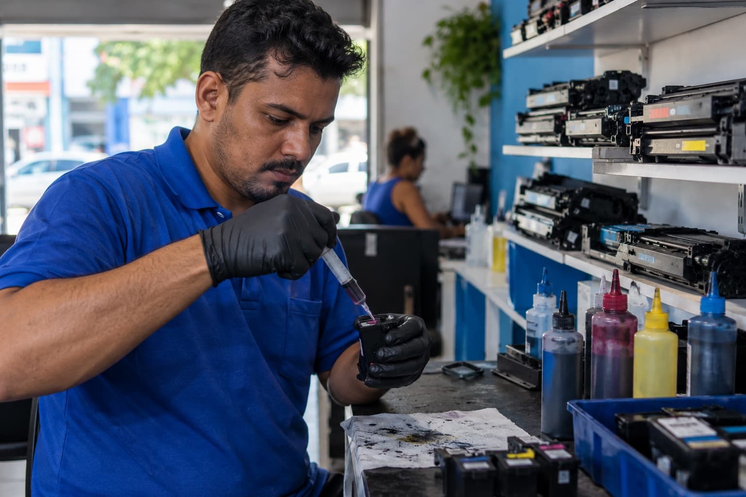Técnico recarregando um cartucho de tinta com seringa na bancada, com toners recarregados na prateleira ao fundo
