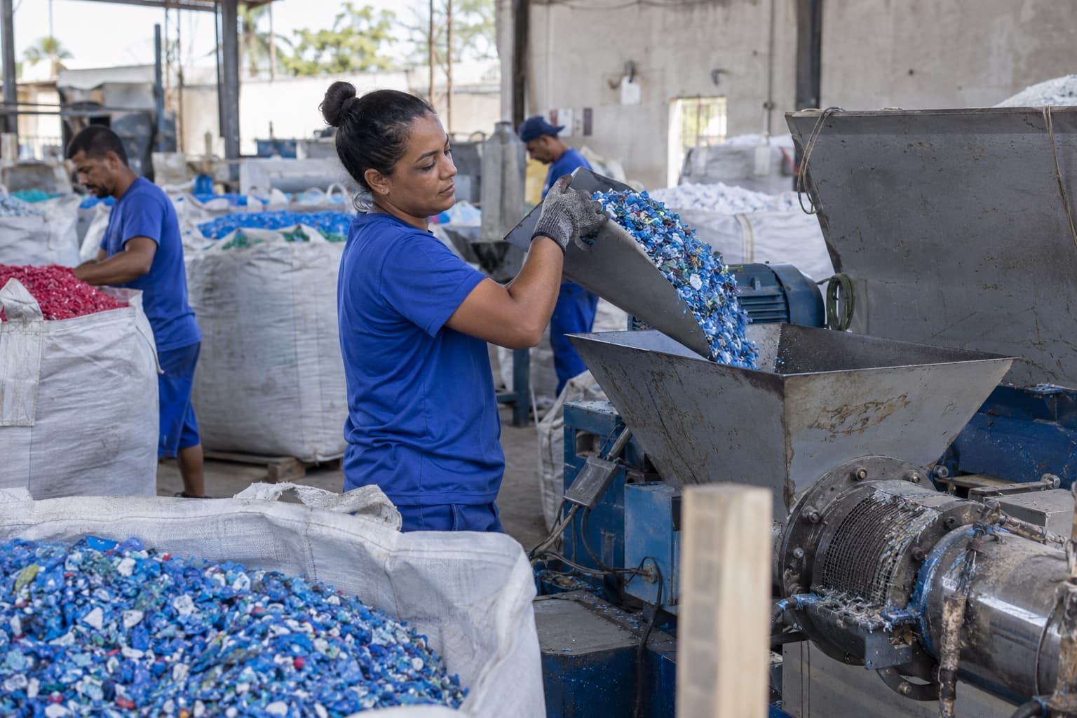 Trabalhadores de uniforme azul despejando flocos de plástico moído no funil de uma máquina numa recicladora com big bags ao redor