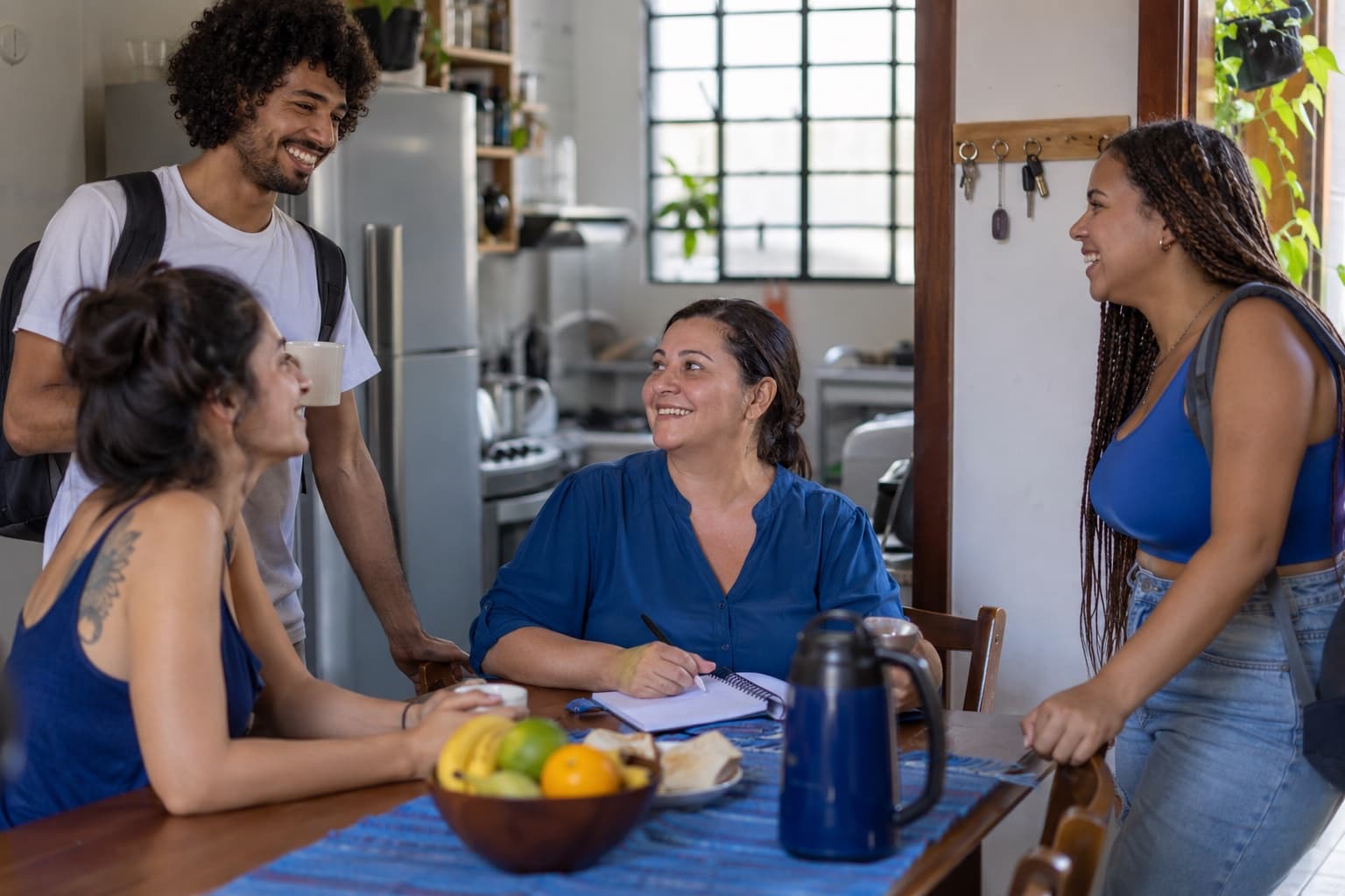 Cozinha de república estudantil com jovens conversando e proprietária de blusa azul com caderno à mesa