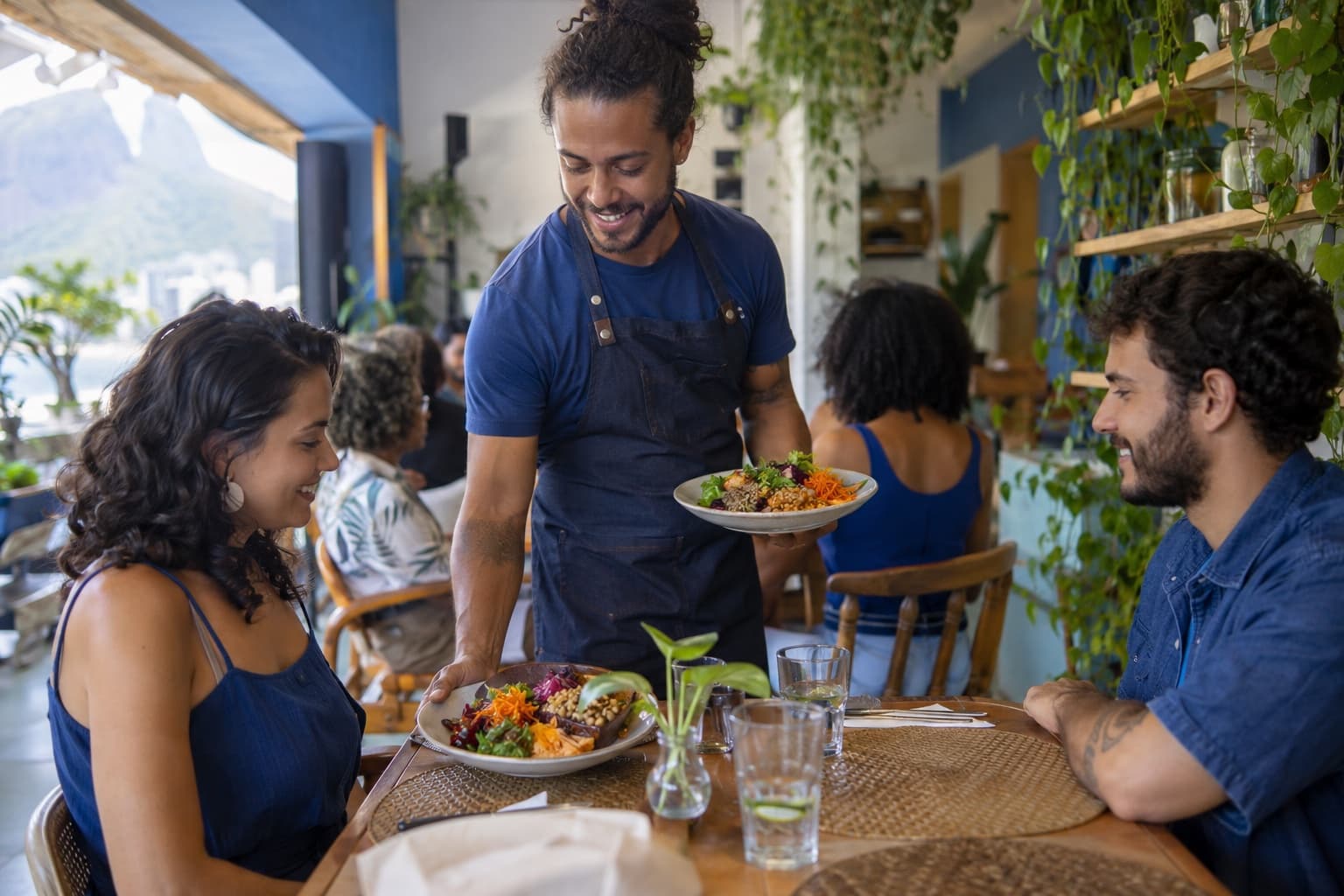 Garçom de camisa azul servindo pratos coloridos com legumes e grãos para clientes em um restaurante vegano claro com plantas nas paredes